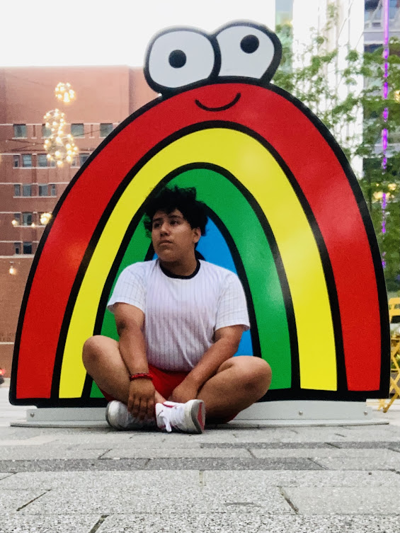 A queer POC with short hair sitting cross-legged in front of a smiling rainbow