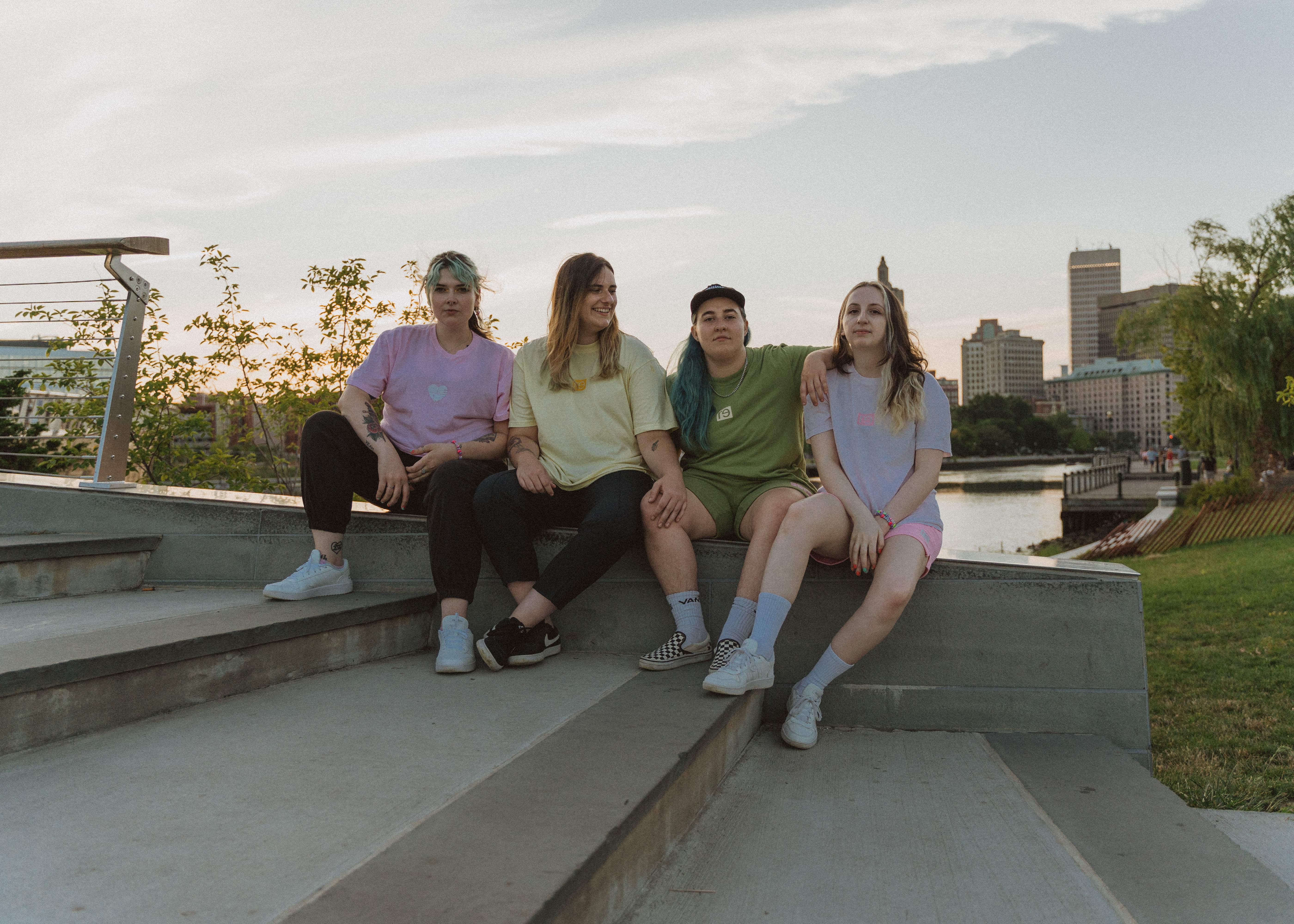 four white people with long hair sit on the ledge along a set of steps with a river and city buildings in the background