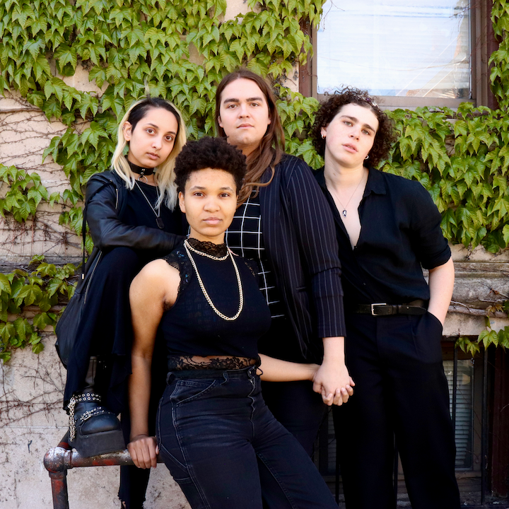4 nonbinary individuals looking at the camera. From left to right: a light brown skinned femme nonbinary person stands with one leg up on a bar. They have shoulder length bleached hair. Next is a lighter skinned black person with natural hair sitting on a pipe/railing and staring at the camera. A long-haired white nonbinary person stands behind them, holding thier hand. Finally a white nonbinary person with chin-length brown curly hair and an open shirt leans into the group. They are all in front of a wall covered in ivy.
