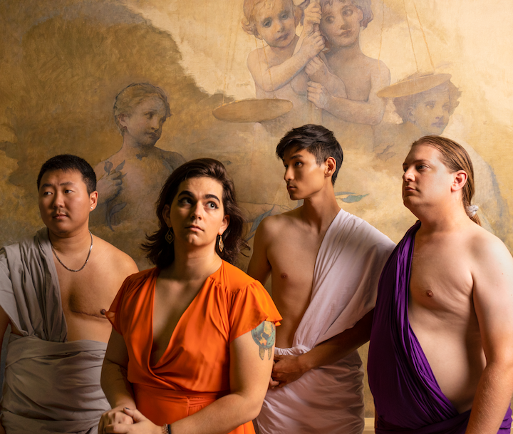 the band wearing togas or similarly flowing clothes, including an asian masculine person with visible top surgery scars, a femme white person in orange with long dark hair, an asian masculine person, and a white masculine person with long blonde hair tied back