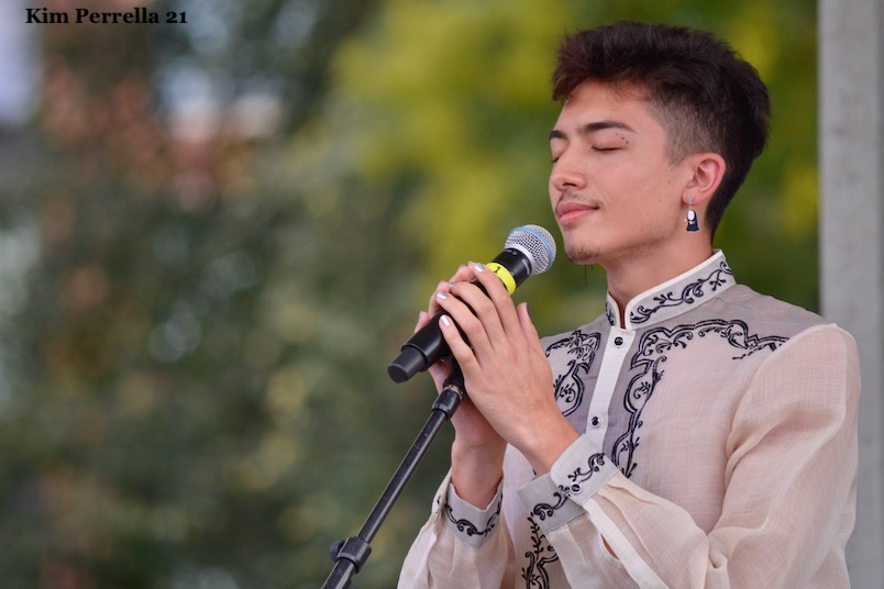 Photo of the Michah Rose performing at Boston Dyke Fest. Michah is a light brown skinned person with short brown hair. Their eyes are closed as they hold a microphone, looking serene. They are wearing a light colored, embroidered shirt that is almost see-through.