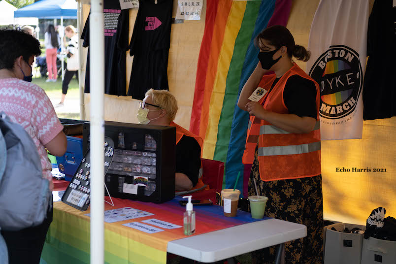 Photo of the Dyke March Merch booth. Raiinbow flags and shirts are hung up in the background. Two masked white/light-skinned people wearing masks are standing behind the table. A white person in a pink and white shirt with short brown hair is looking at the Merch on the table.