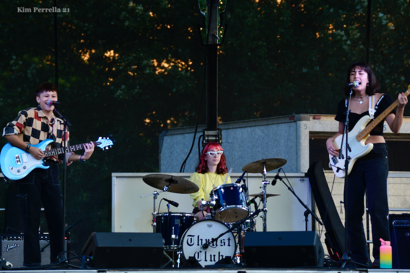 Photo of Addie. Left is an asian person with short dark hair wearing a tan shirt with black and red squares playing a blue bass. Center is a white person in a yellow shirt with red hair and white glasses playing drums. Right is a light skinned person with medium length dark hair singing and playing a white guitar.