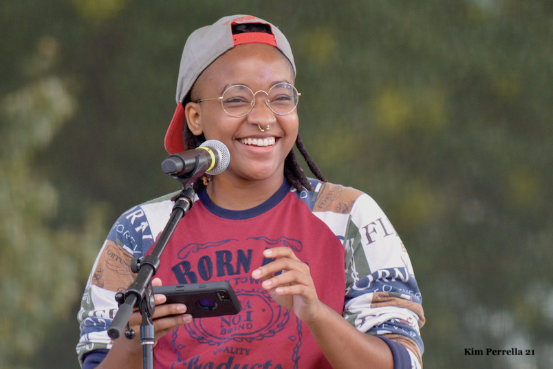 Photo of Max McLune. A smiling black person with shoulder-length dredlocks wearing a backwards grey hat, a mostly red shirt, and round glasses. The person is holding a phone and smiling.